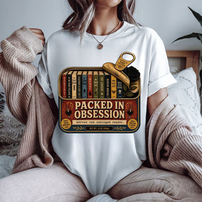 A person wearing a white graphic T-shirt sits on a bed with a beige knit sweater draped off the shoulders. The shirt features a vintage-style book tin graphic with the text 'PACKED IN OBSESSION' and illustrated books, a gold key, and a black rose.