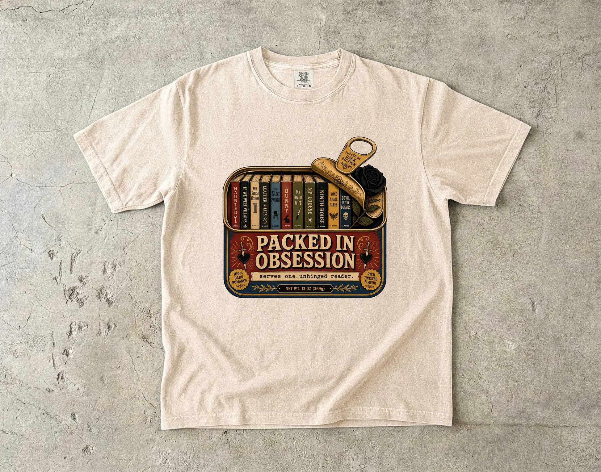 A cream-colored short-sleeve t-shirt with a vintage-style graphic on the front reading 'PACKED IN OBSESSION' over a bookshelf and a magnifying glass, laid flat on a textured gray concrete surface.