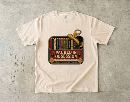 A cream-colored short-sleeve t-shirt with a vintage-style graphic on the front reading 'PACKED IN OBSESSION' over a bookshelf and a magnifying glass, laid flat on a textured gray concrete surface.