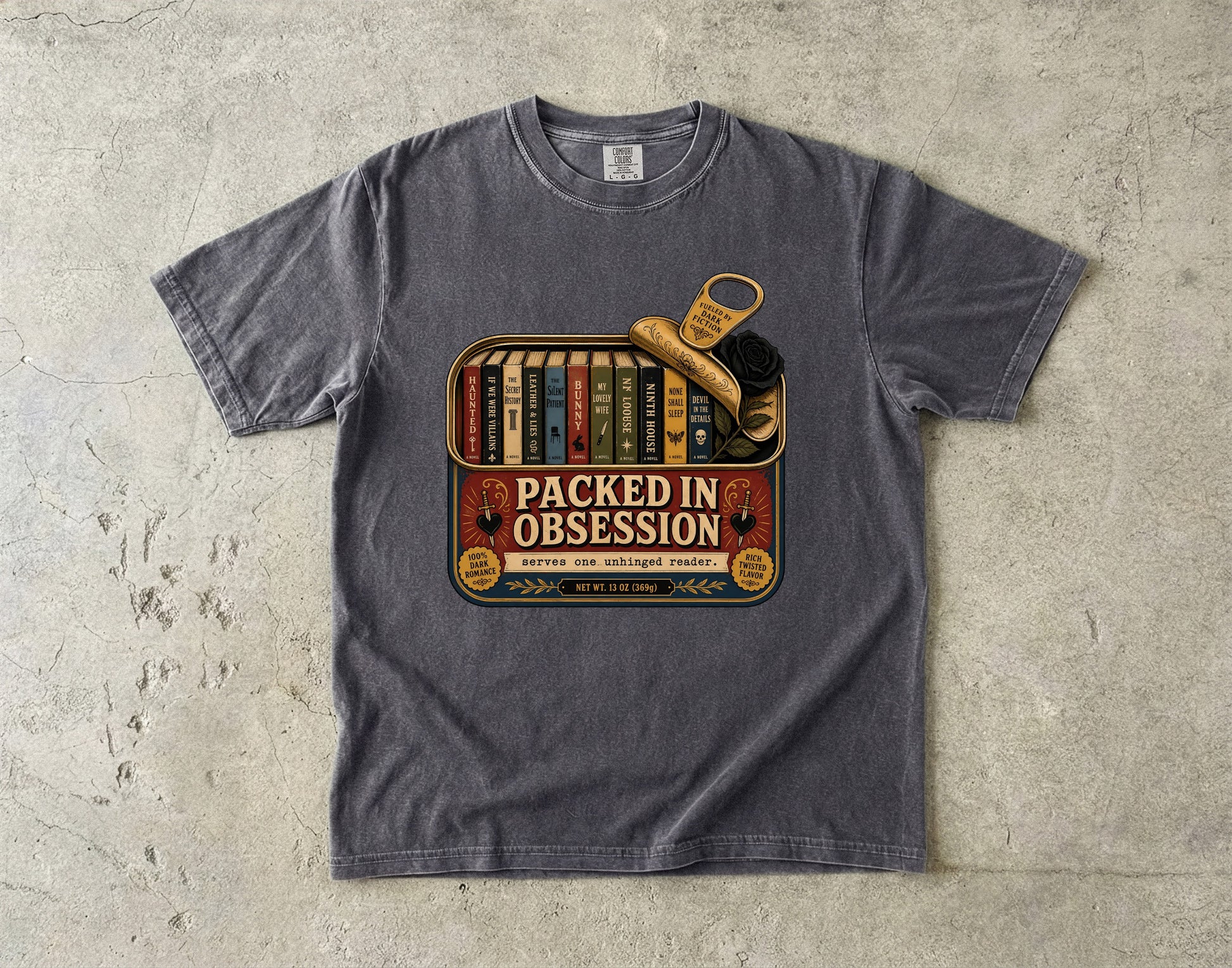 A dark gray heather t-shirt laid flat on a textured concrete surface, featuring a vintage-style graphic print with the text 'PACKED IN OBSESSION' and illustrations of books, a key, and a shoe.