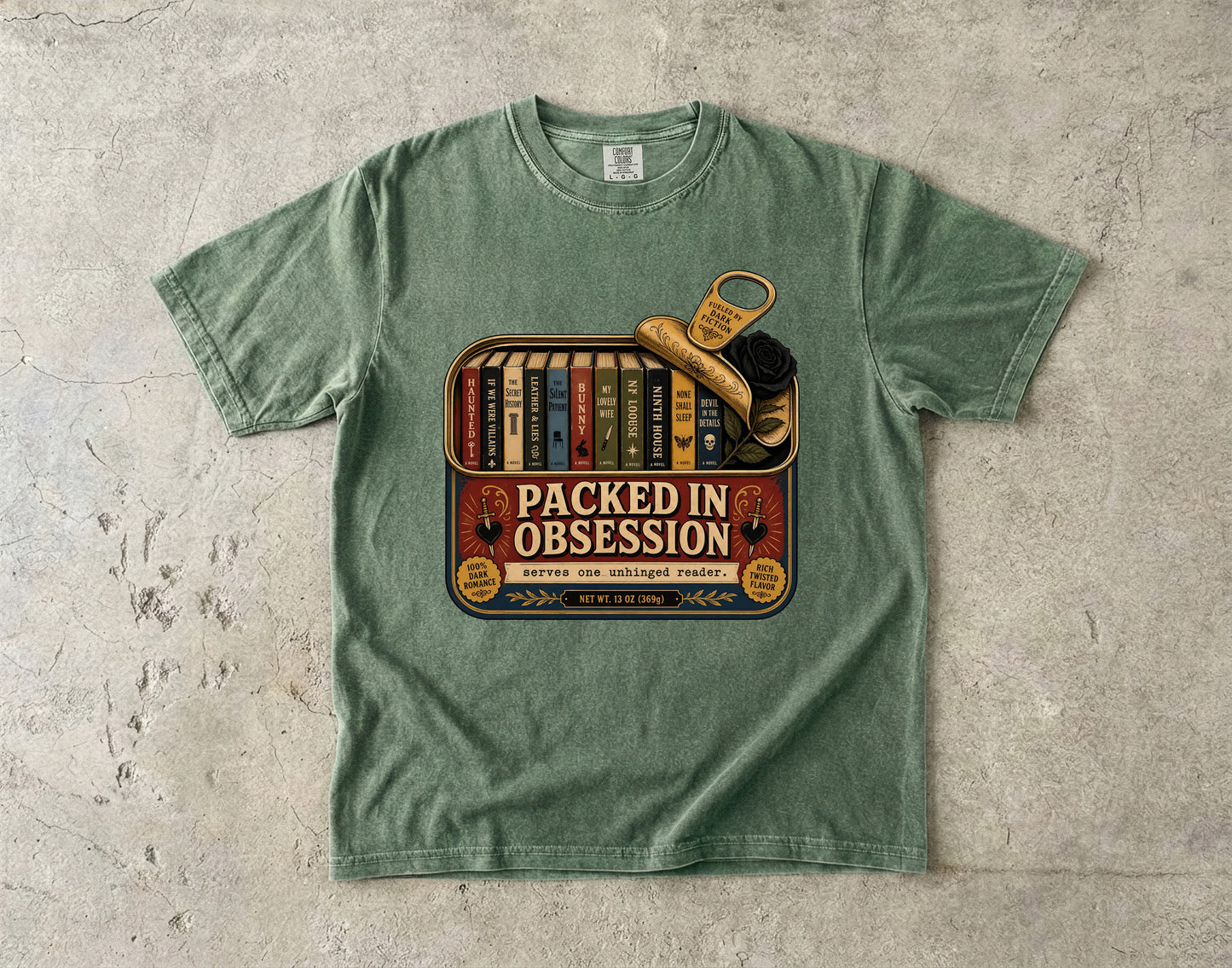 A sage green crewneck t-shirt laid flat on a textured concrete surface, featuring a vintage-style graphic print of a bookshelf filled with books and a leather boot with a keychain, labeled 'PACKED IN OBSESSION'.