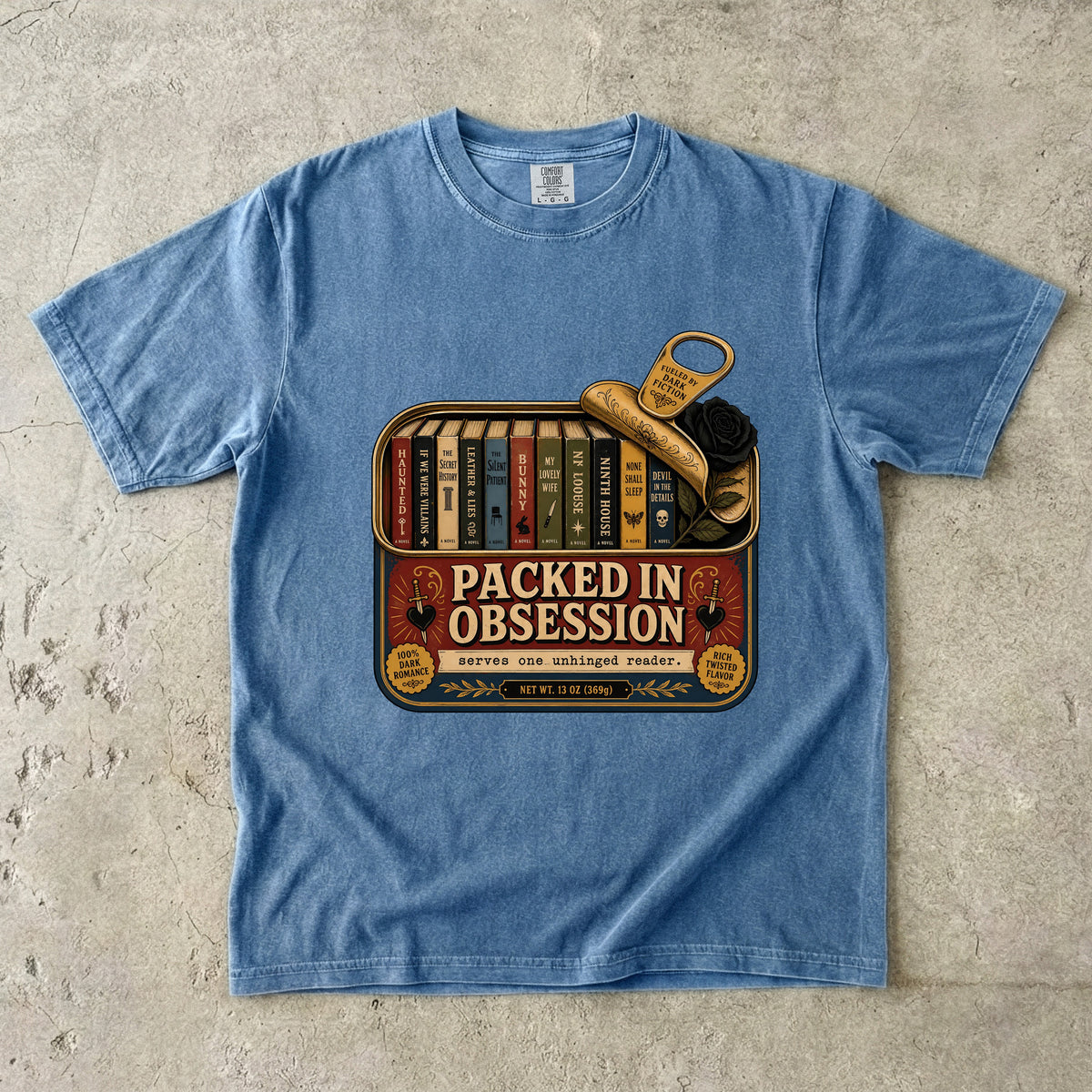 A light blue crewneck t-shirt laid flat on a textured concrete surface, featuring a vintage-style graphic of a bookshelf filled with books and a gold key, with the text 'PACKED IN OBSESSION'.