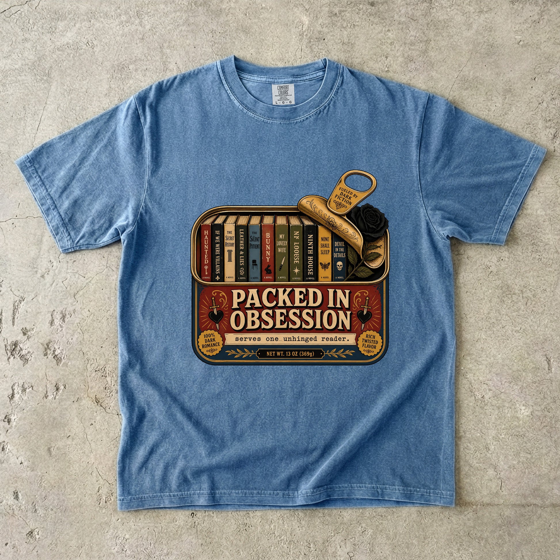 A light blue crewneck t-shirt laid flat on a textured concrete surface, featuring a vintage-style graphic of a bookshelf filled with books and a gold key, with the text 'PACKED IN OBSESSION'.