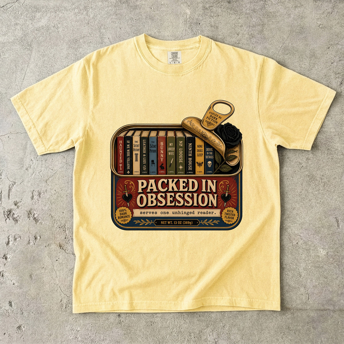 A pale yellow short-sleeve t-shirt laid flat on a textured gray concrete surface. The front of the shirt features a large graphic print depicting a vintage bookshelf filled with books, a pair of gold scissors, and a black rose, with the text 'PACKED IN OBSESSION' and 'serve and observe product'.