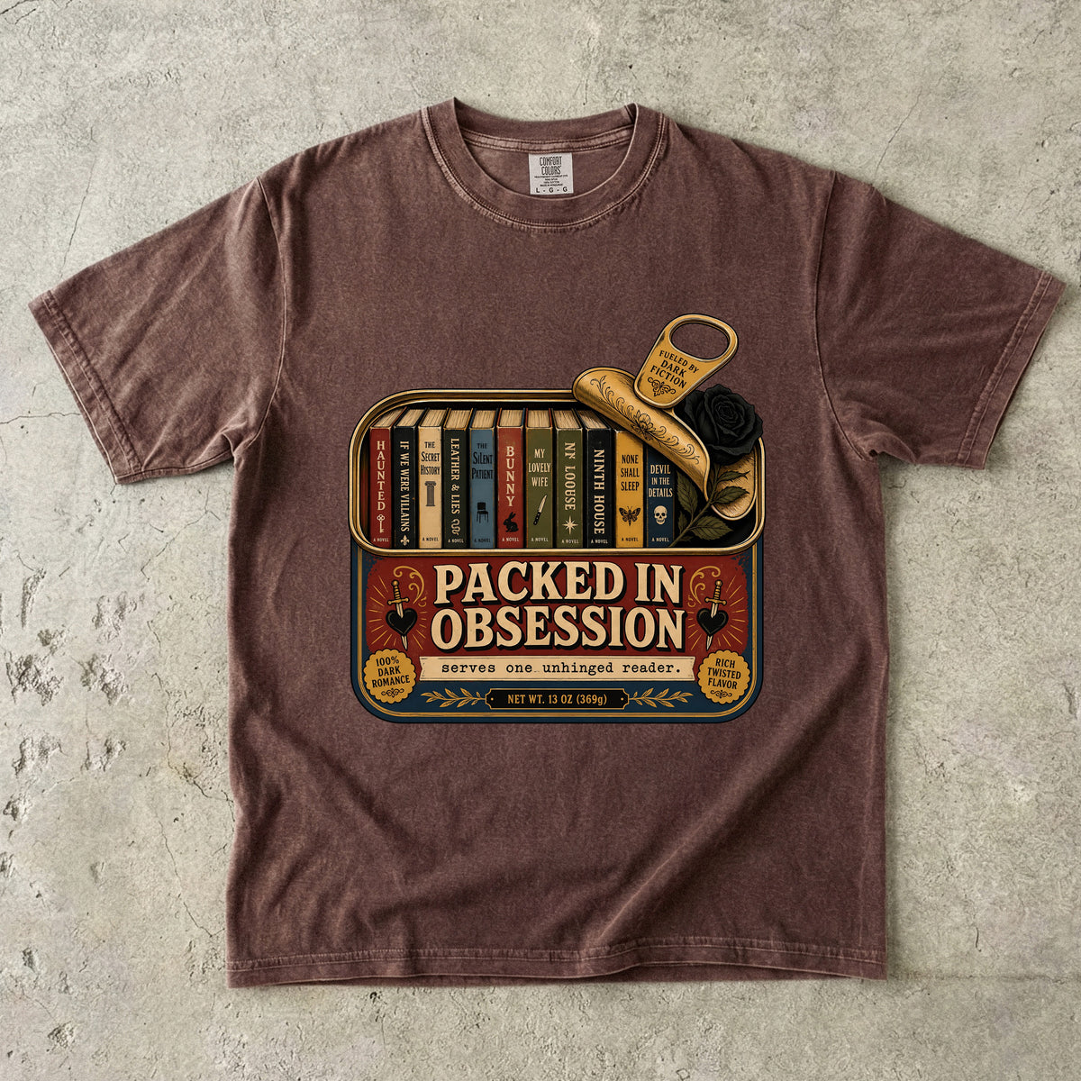 A brown short-sleeve crewneck t-shirt laid flat on a textured gray concrete surface. The shirt features a vintage-style graphic print of a book tin with the text 'PACKED IN OBSESSIVE' and illustrations of books, a key, and a rose.