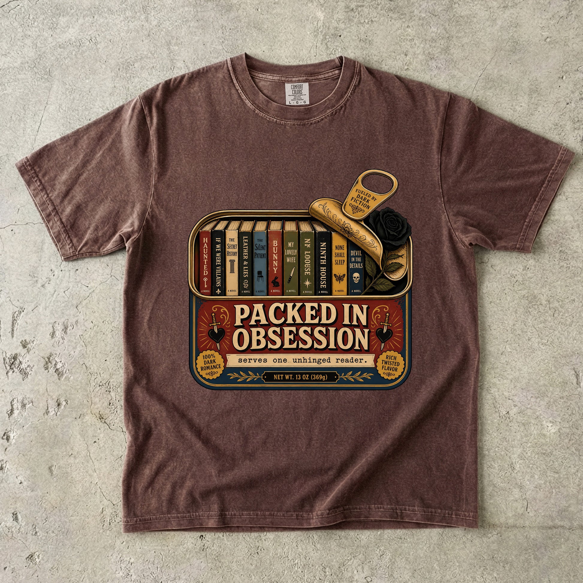 A brown short-sleeve crewneck t-shirt laid flat on a textured gray concrete surface. The shirt features a vintage-style graphic print of a book tin with the text 'PACKED IN OBSESSIVE' and illustrations of books, a key, and a rose.