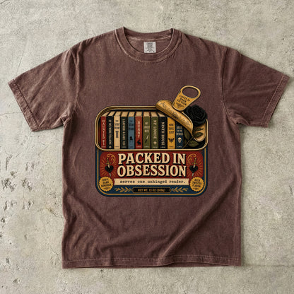 A brown short-sleeve crewneck t-shirt laid flat on a textured gray concrete surface. The shirt features a vintage-style graphic print of a book tin with the text 'PACKED IN OBSESSIVE' and illustrations of books, a key, and a rose.