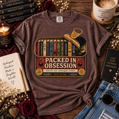 A brown short-sleeve graphic T-shirt laid flat on a wooden surface, styled with books, a candle, a mug, sunglasses, and denim. The shirt has a large vintage-style book spine graphic and the text 'PACKED IN OBSESSION' with smaller slogan text below.