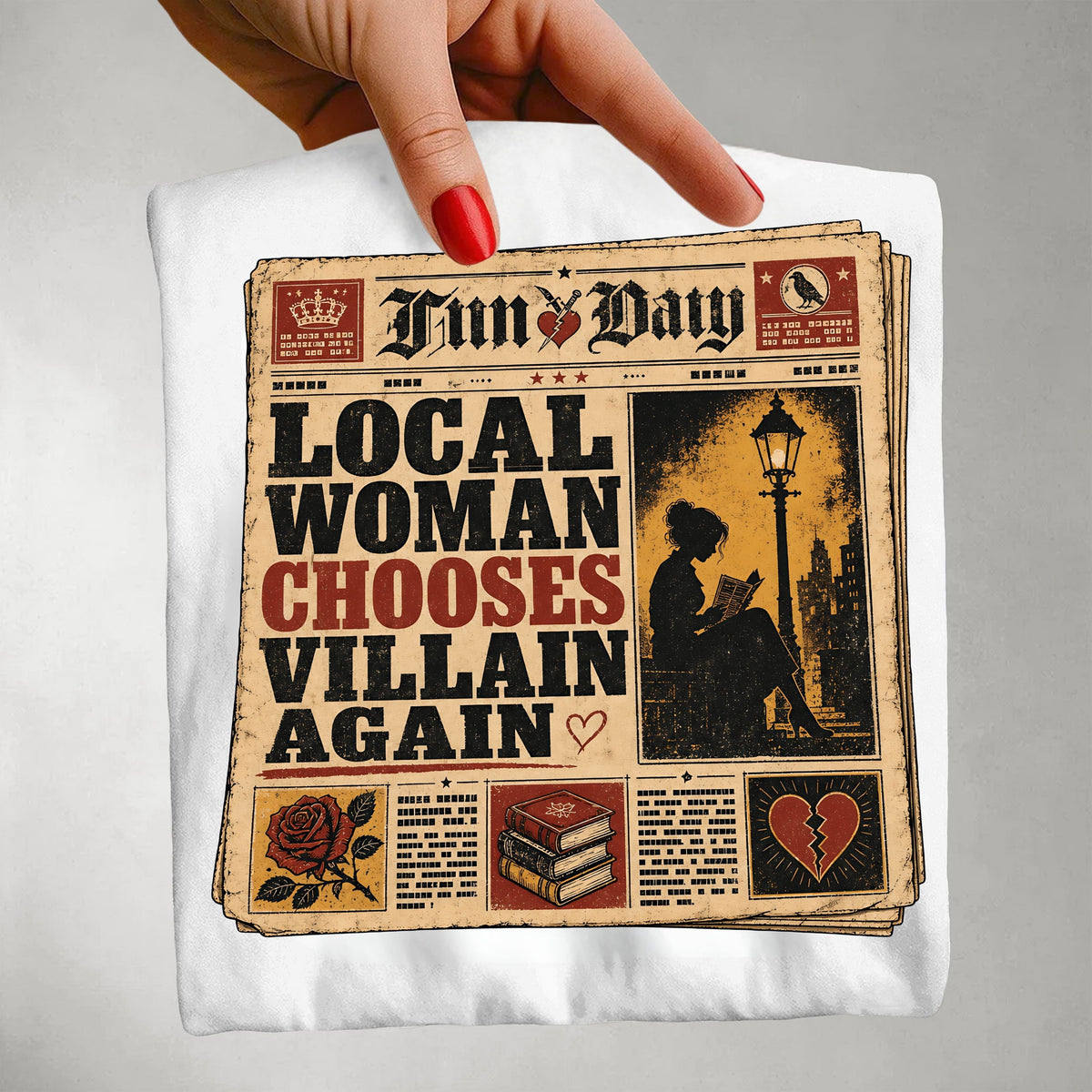A hand with red nail polish holds up a white T-shirt featuring a vintage newspaper graphic with the text 'LOCAL WOMAN CHOOSES VILLAIN AGAIN' and a sepia-toned illustration of a woman reading under a streetlamp.