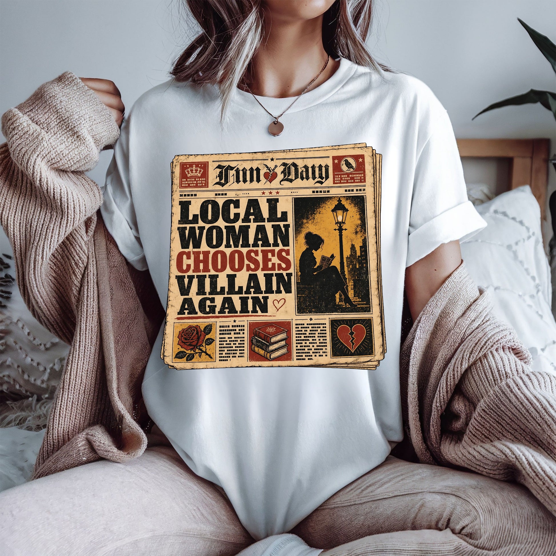 A woman wearing a white t-shirt with a vintage newspaper graphic that reads 'LOCAL WOMAN CHOOSES VILLAIN AGAIN' under the header 'Fun Day'. She is sitting in a cozy room with a beige knit sweater draped off her shoulders.