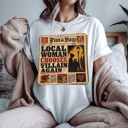 A woman wearing a white t-shirt with a vintage newspaper graphic that reads 'LOCAL WOMAN CHOOSES VILLAIN AGAIN' under the header 'Fun Day'. She is sitting in a cozy room with a beige knit sweater draped off her shoulders.