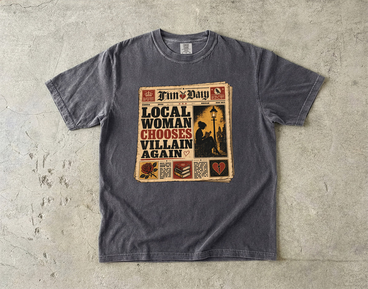 A dark gray crewneck t-shirt laid flat on a textured concrete surface, featuring a vintage newspaper graphic with the text 'Local Woman Chooses Villain Again' and 'Fun & Day'.