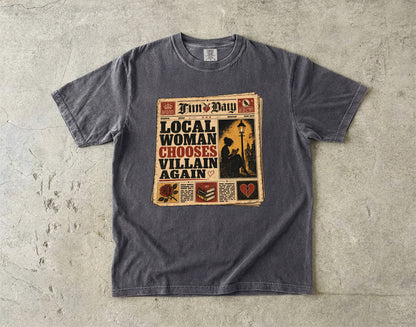 A dark gray crewneck t-shirt laid flat on a textured concrete surface, featuring a vintage newspaper graphic with the text 'Local Woman Chooses Villain Again' and 'Fun & Day'.