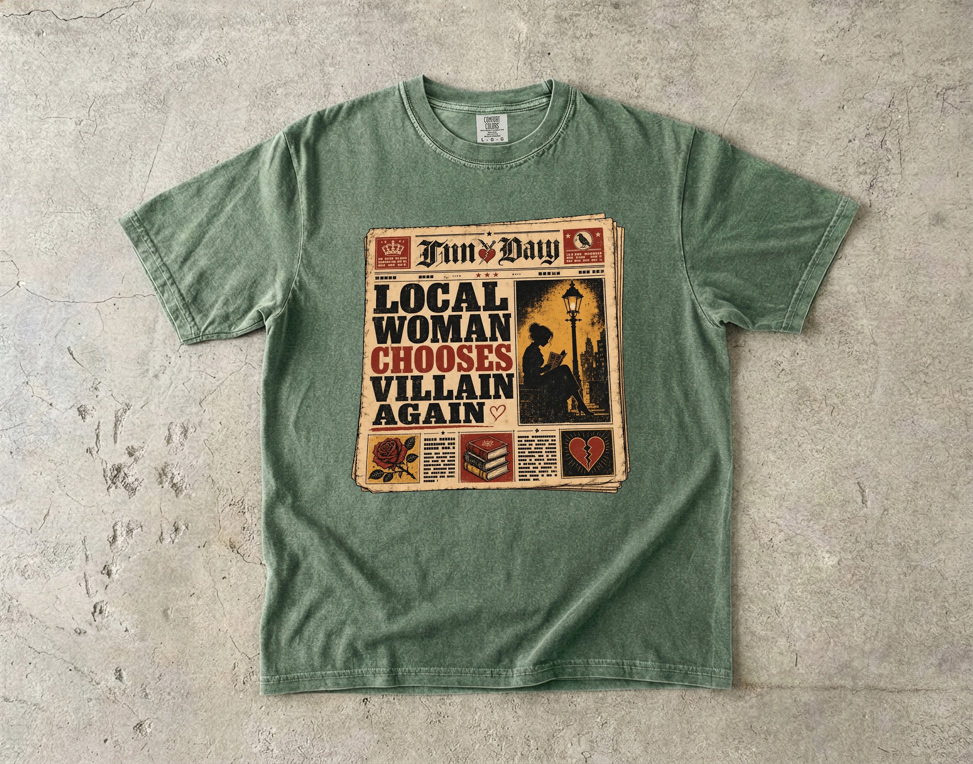 A sage green crewneck t-shirt laid flat on a textured concrete surface, featuring a vintage newspaper graphic with the text 'LOCAL WOMAN CHOOSES VILLAIN AGAIN' and 'Fun's Day'.