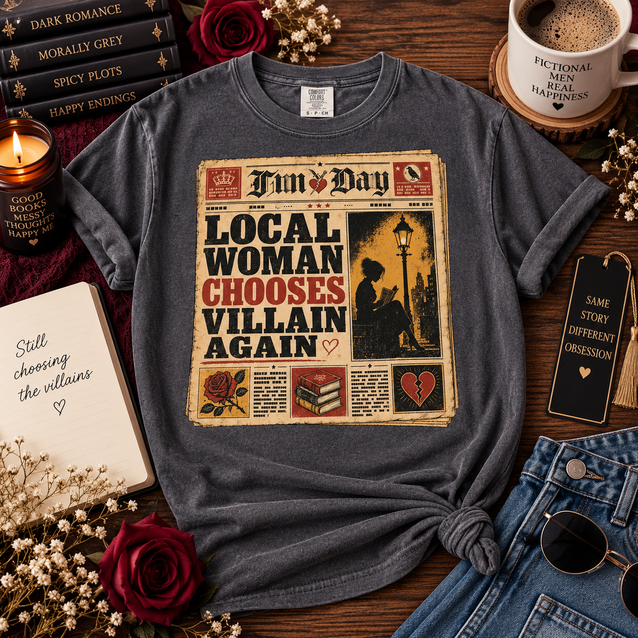 A dark heather gray graphic t-shirt styled in a cozy flat-lay arrangement on a wooden table, surrounded by books, a candle, a coffee cup, a notebook, sunglasses, and red roses. The t-shirt features a newspaper-style graphic with the text 'Fun Day LOCAL WOMAN CHOOSES VILLAIN AGAIN'.