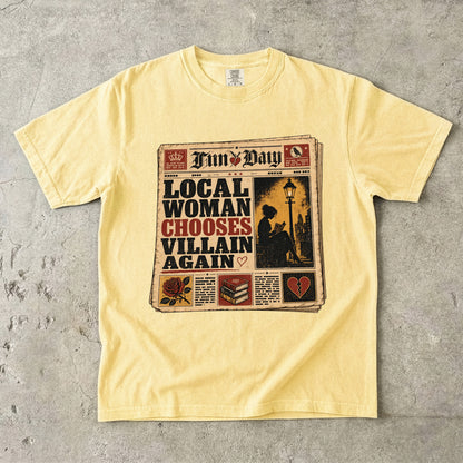 A pale yellow short-sleeve t-shirt laid flat on a textured gray concrete surface. The front of the shirt features a large graphic print styled like a newspaper front page with the text 'LOCAL WOMAN CHOOSES VILLAIN AGAIN' and the title 'Fun Day'.