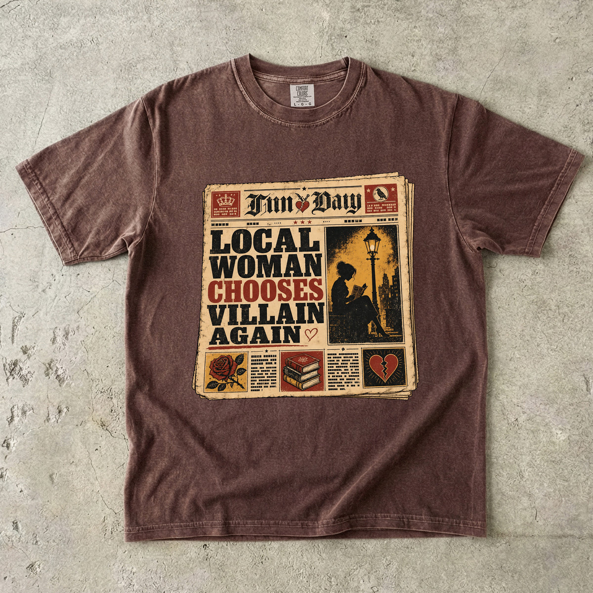 A washed brown short-sleeve t-shirt laid flat on a textured gray concrete surface, featuring a vintage newspaper graphic on the front with the headline 'LOCAL WOMAN CHOOSES VILLAIN AGAIN'.