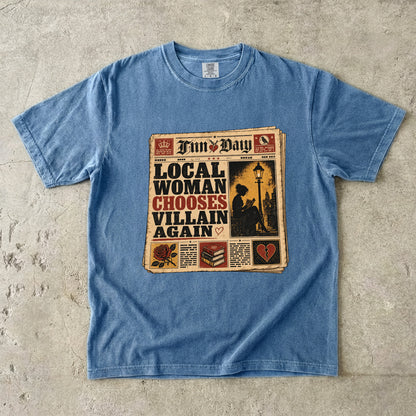 A light blue short-sleeve t-shirt laid flat on a textured concrete surface, featuring a vintage newspaper graphic on the front with the headline 'LOCAL WOMAN CHOOSES VILLAIN AGAIN' and a newspaper masthead 'Tim's Day'.