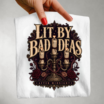 A hand with red nail polish holds up a white t-shirt featuring a gothic-style graphic print that reads 'LIT BY BAD IDEAS AFTER MIDNIGHT' around an illustration of three lit candles, roses, and ornate filigree.