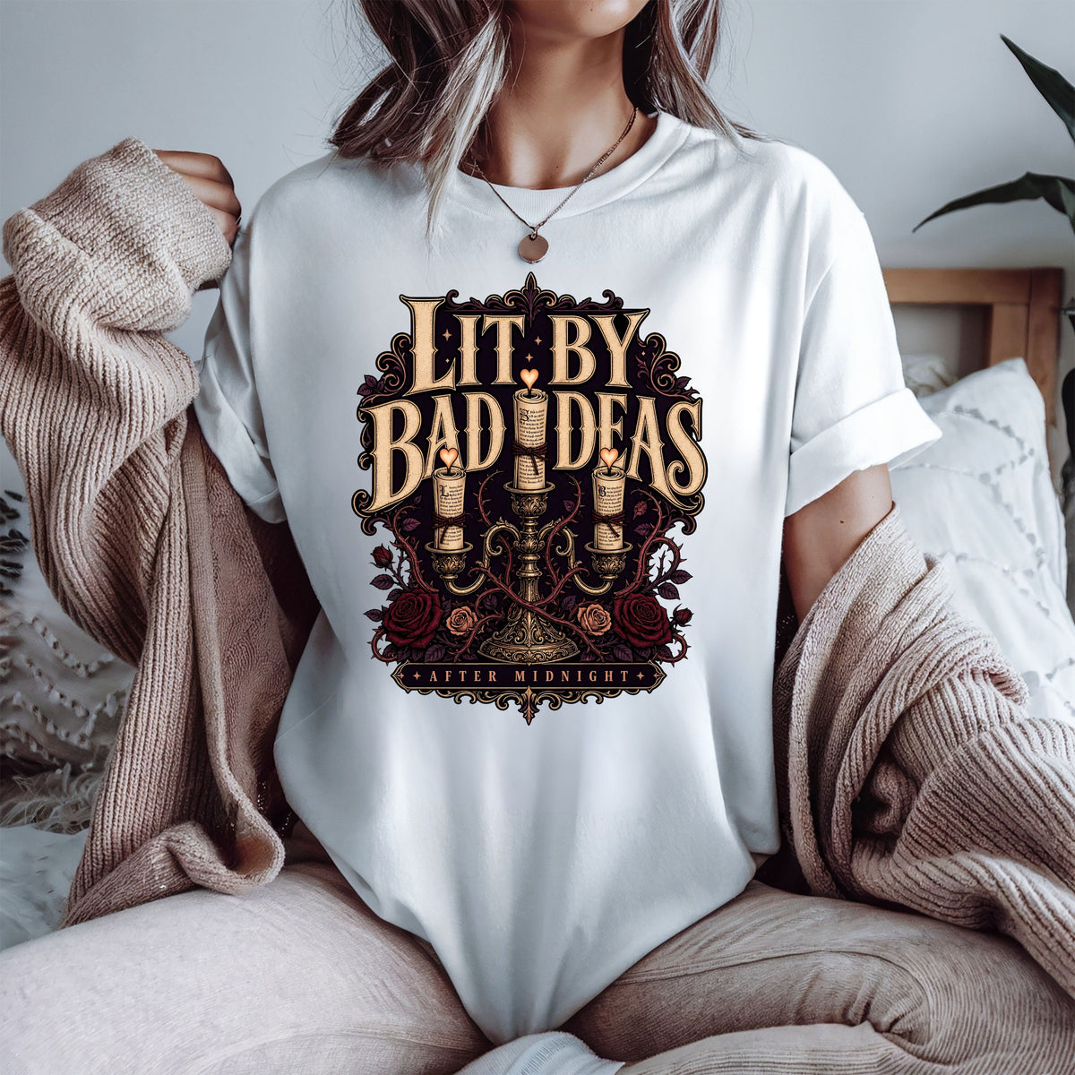 A person wearing a white graphic T-shirt with a large vintage-style black and gold print reading 'Lit. By Bad Ideas' and 'After Midnight,' styled with a beige knit sweater draped off the shoulders in a cozy indoor setting.