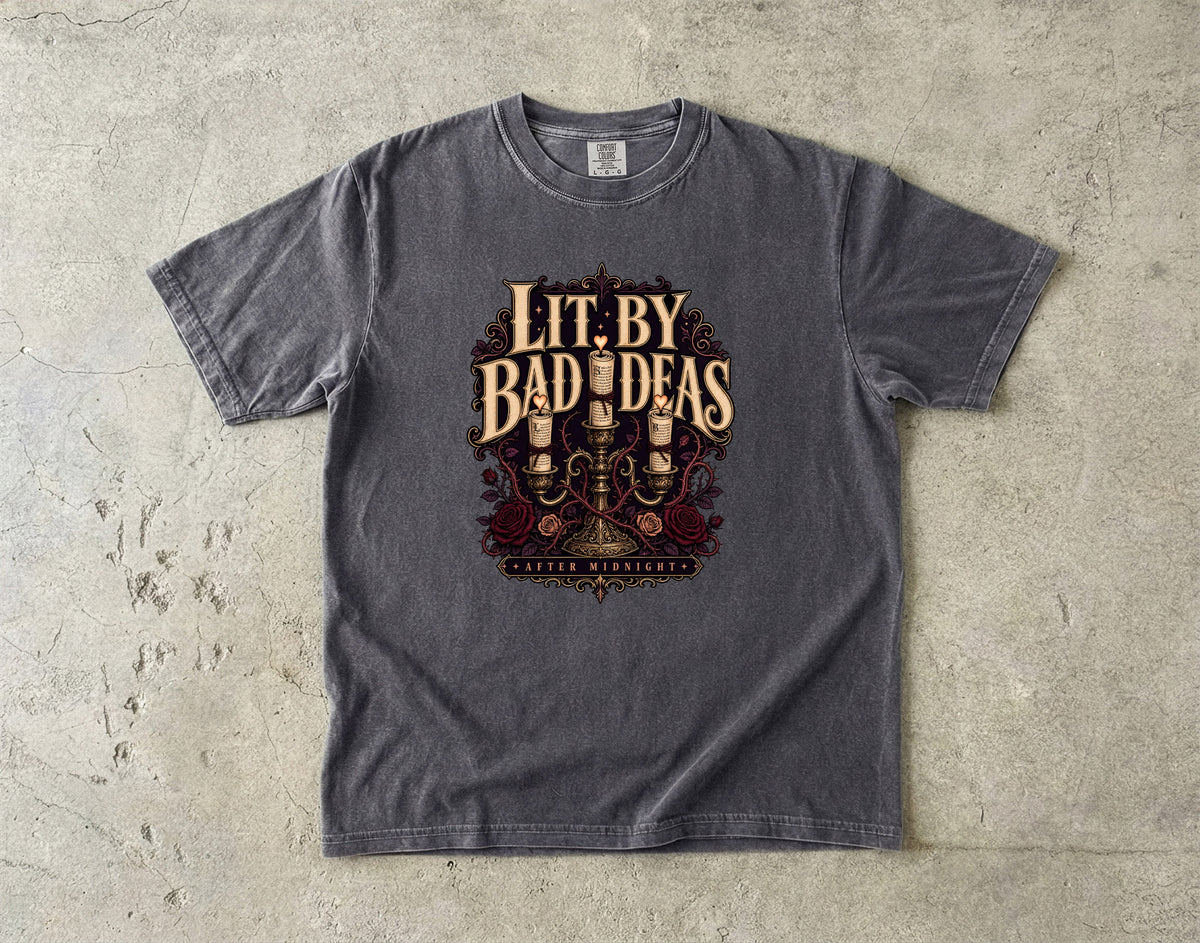 A dark gray heather t-shirt with a vintage-style graphic reading 'Lit. By Bad Ideas' and 'Western Westerners' beneath it, laid flat on a textured concrete surface.
