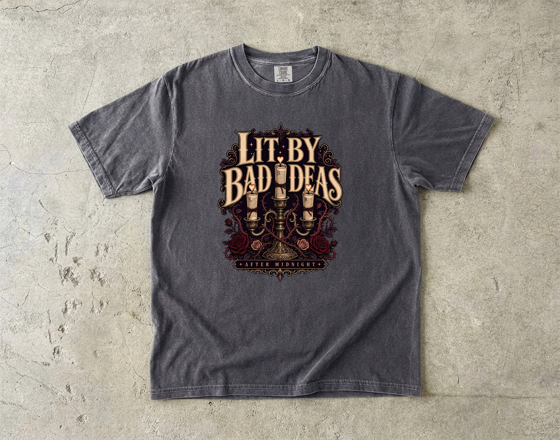 A dark gray heather t-shirt with a vintage-style graphic reading 'Lit. By Bad Ideas' and 'Western Westerners' beneath it, laid flat on a textured concrete surface.