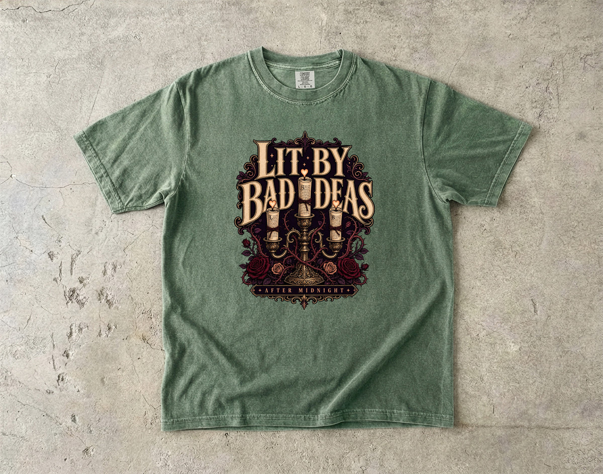 A sage green crewneck t-shirt laid flat on a textured concrete surface, featuring a vintage-style graphic print with the text 'Lit. By Bad Ideas' and 'Guilty Men & Women' in a dark red and gold illustration.