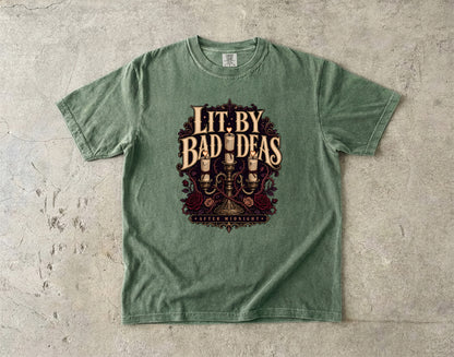 A sage green crewneck t-shirt laid flat on a textured concrete surface, featuring a vintage-style graphic print with the text 'Lit. By Bad Ideas' and 'Guilty Men & Women' in a dark red and gold illustration.