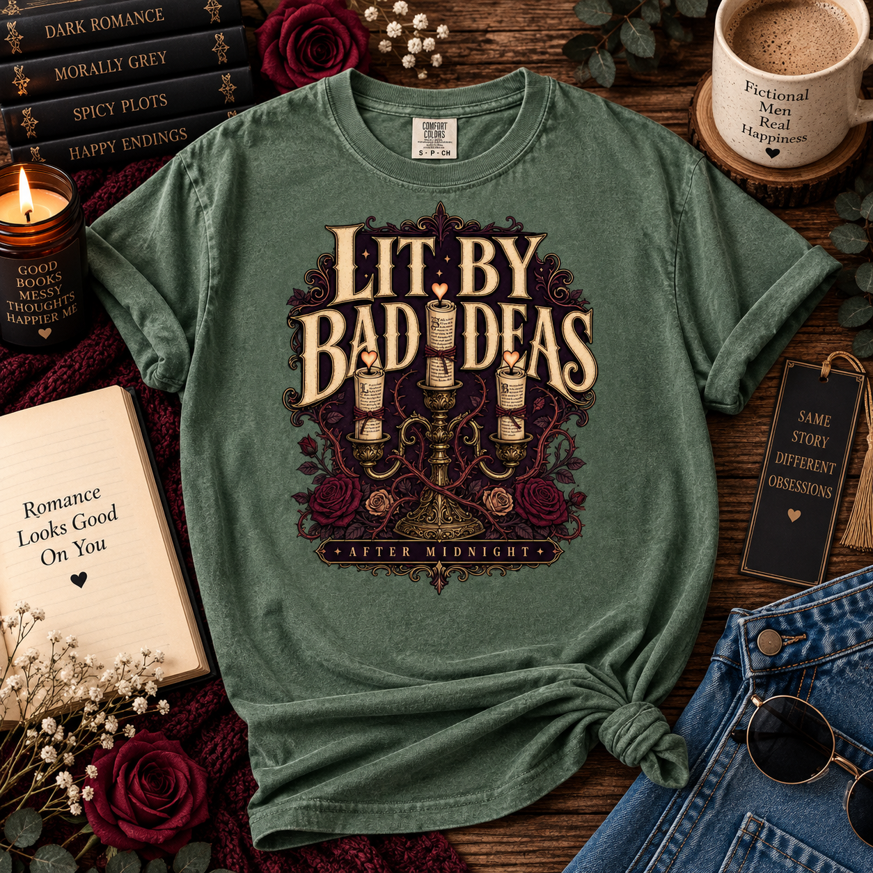 A green graphic t-shirt styled in a cozy flat-lay arrangement with books, a candle, a coffee mug, jeans, and sunglasses on a wooden surface. The shirt features a gothic graphic that reads 'Lit by Bad Ideas' with a candlestick and roses.