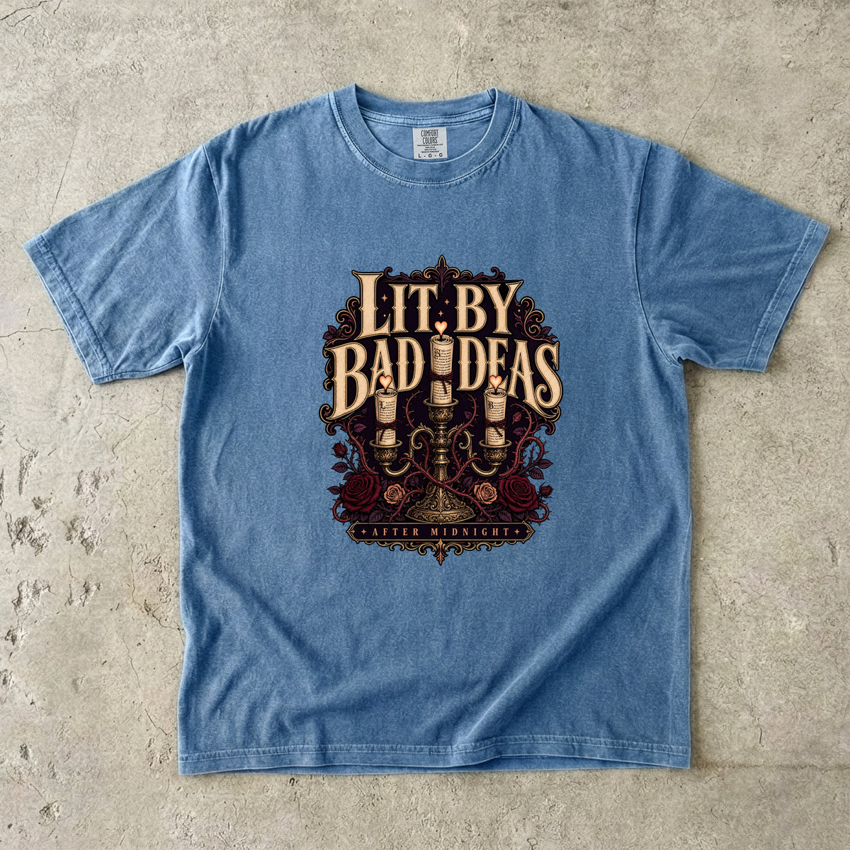 A light blue short-sleeve t-shirt with a vintage-style graphic on the front reading 'LIT. BY BAD IDEAS' and 'GODDEYS MANSION', laid flat on a textured concrete surface.