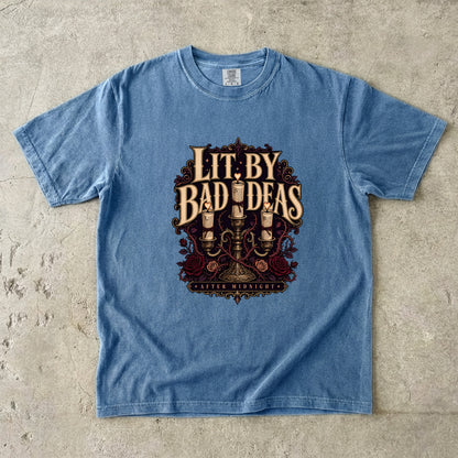A light blue short-sleeve t-shirt with a vintage-style graphic on the front reading 'LIT. BY BAD IDEAS' and 'GODDEYS MANSION', laid flat on a textured concrete surface.