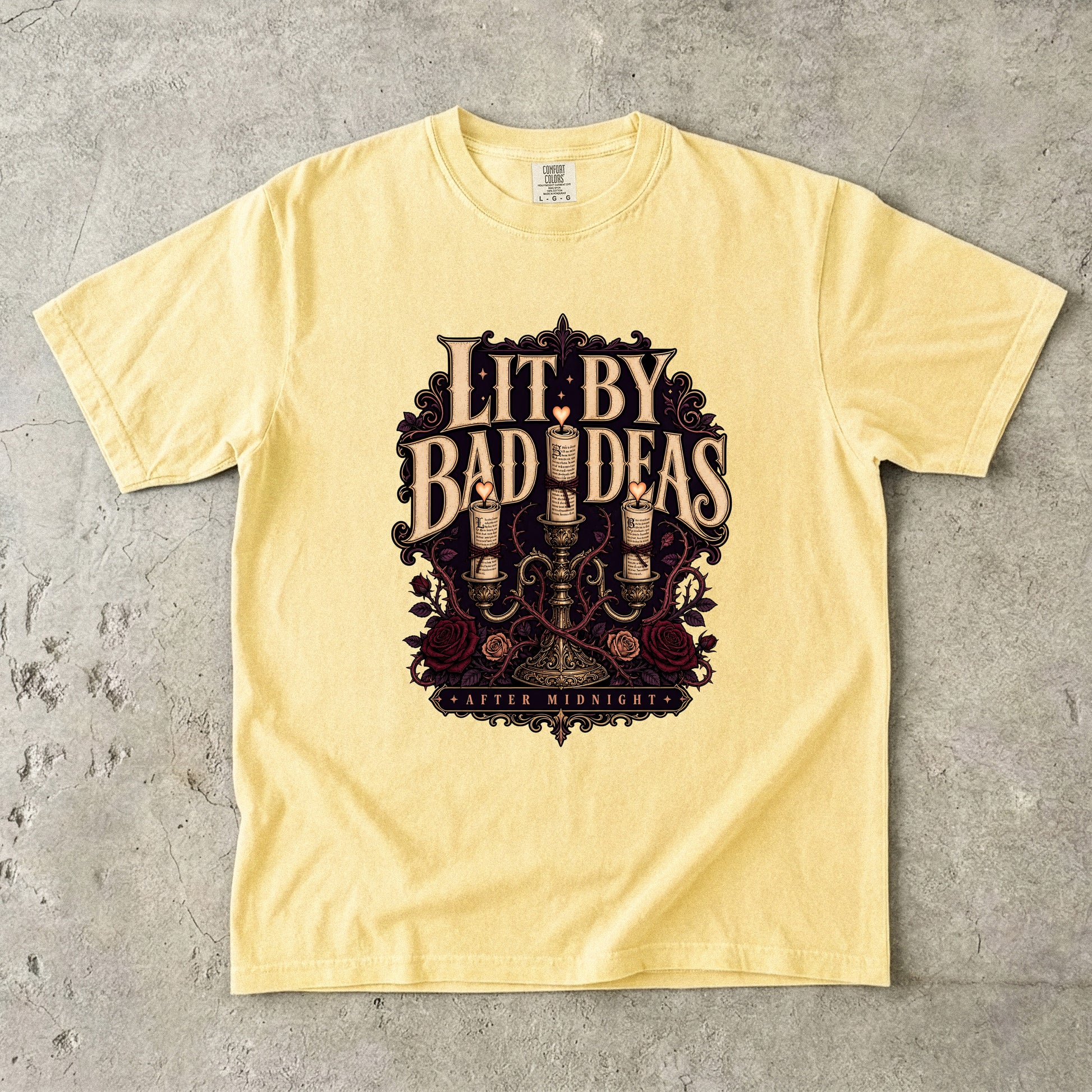 A pale yellow graphic t-shirt laid flat on a textured gray concrete surface. The shirt features a large, ornate graphic in the center with the text 'LIT.
