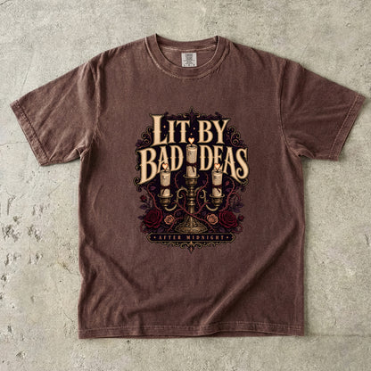 A brown short-sleeve graphic t-shirt laid flat on a textured gray concrete background. The shirt features a vintage-style graphic with the text 'LIT BY BAD IDEAS' in a serif font, decorated with illustrations of candles, roses, and a chandelier, and 'MATTER MIDNIGHT' at the bottom.