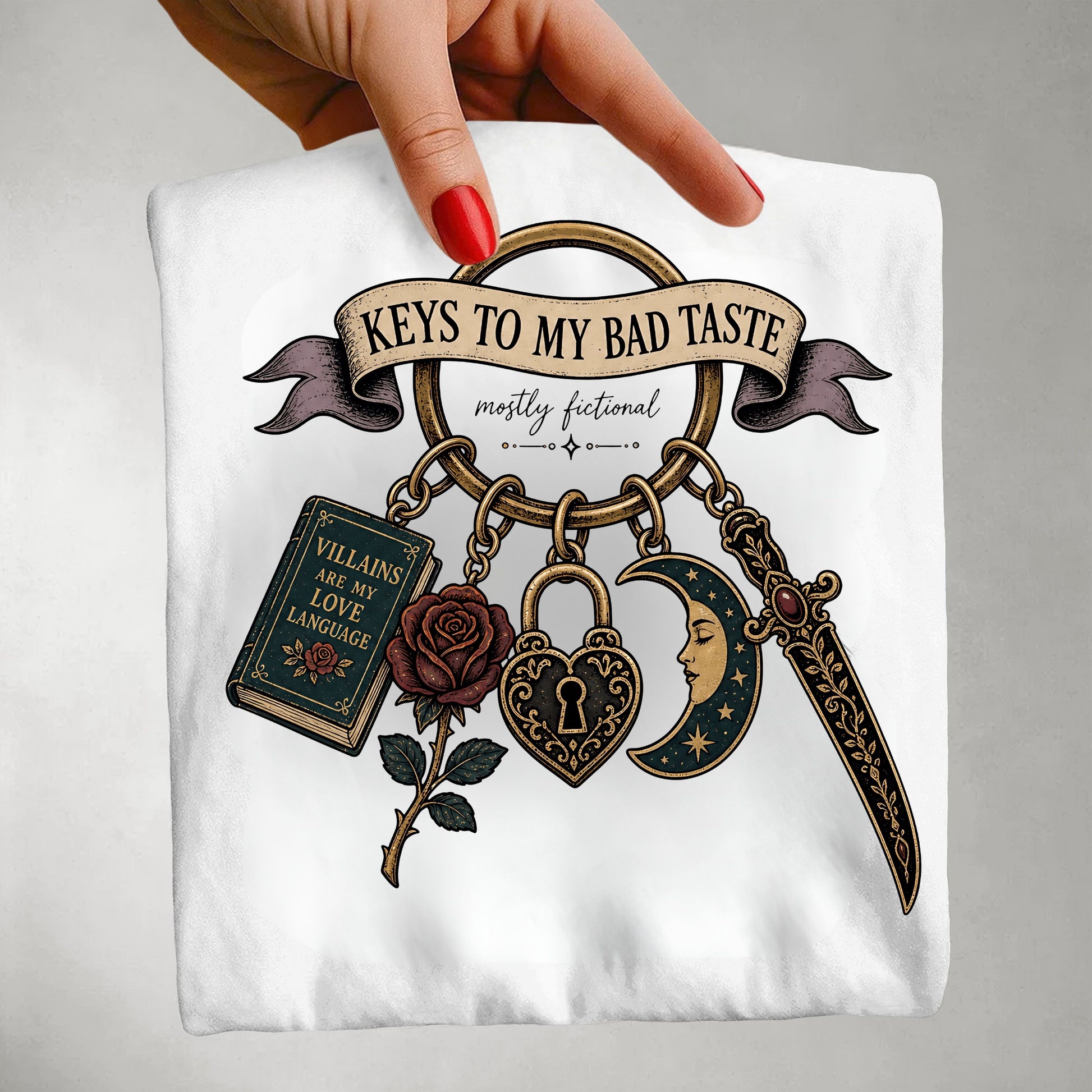 A hand with red nail polish holds up a white t-shirt featuring a detailed graphic print. The graphic shows a keyring with a rose, padlock, dagger, book, and crescent moon under a banner that reads 'KEYS TO MY BAD TASTE mostly fictional'.