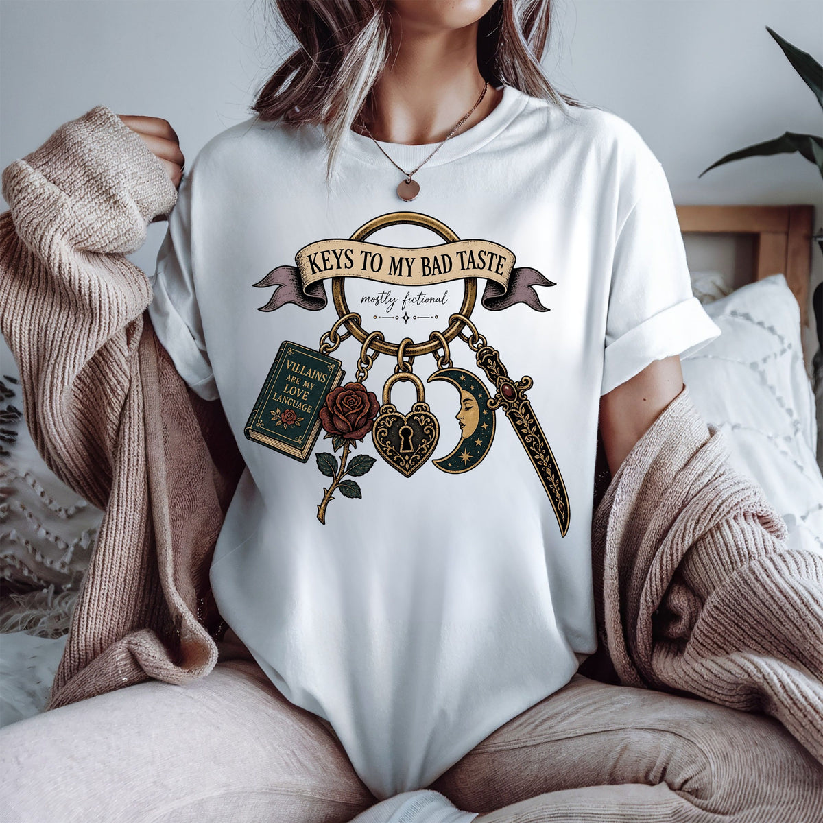 A woman wearing a white graphic t-shirt with a gothic-themed design that reads 'Keys to My Bad Taste' and features a keychain with a book, rose, heart, dagger, and crescent moon. She is sitting in a cozy setting with a beige knit sweater draped over her shoulders.