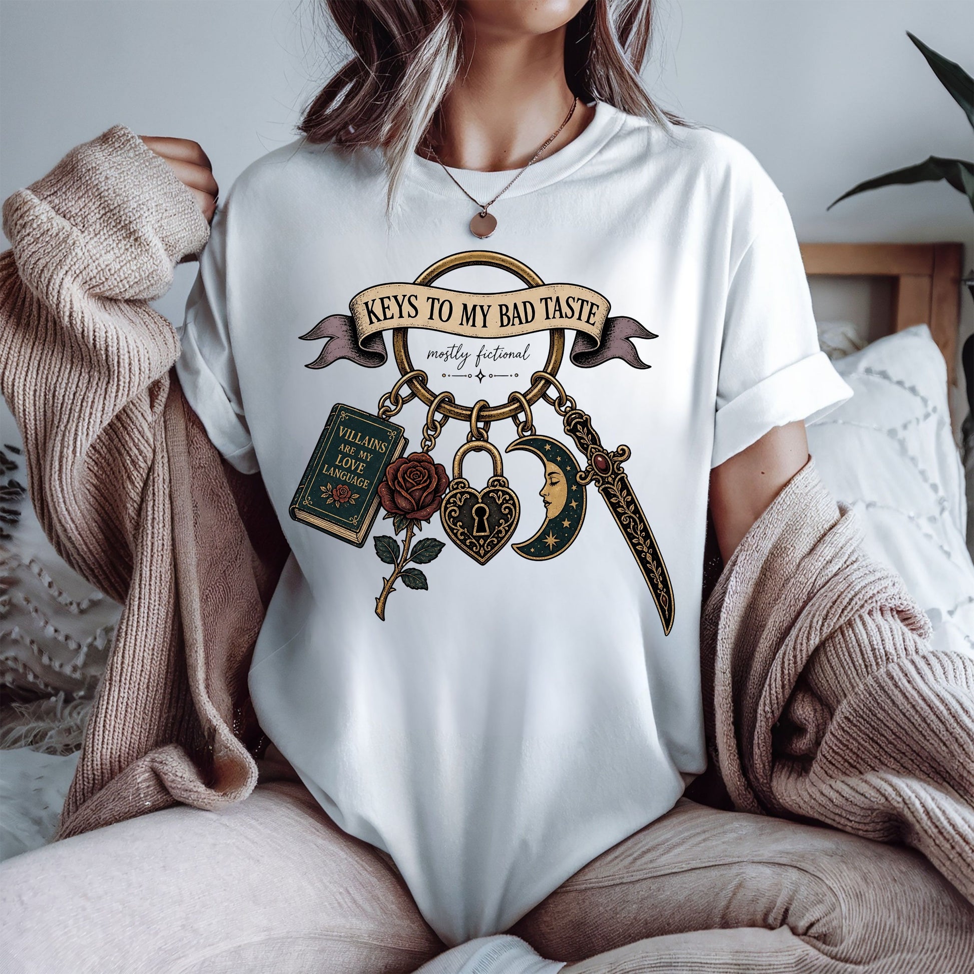 A woman wearing a white graphic t-shirt with a gothic-themed design that reads 'Keys to My Bad Taste' and features a keychain with a book, rose, heart, dagger, and crescent moon. She is sitting in a cozy setting with a beige knit sweater draped over her shoulders.