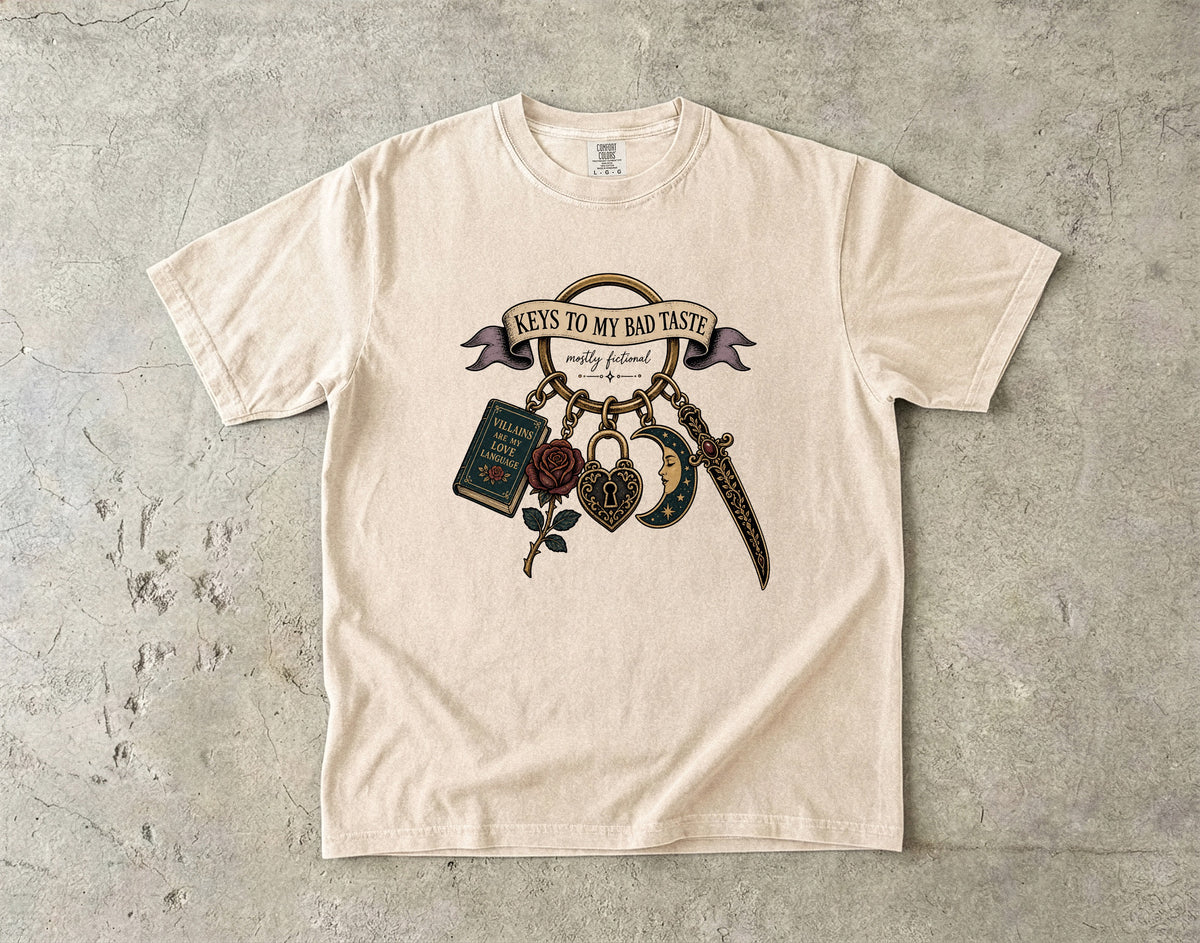 A cream-colored short-sleeve t-shirt with a graphic print of 'Keys to My Bad Taste' hanging keys, laid flat on a textured gray concrete surface.