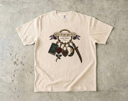 A cream-colored short-sleeve t-shirt with a graphic print of 'Keys to My Bad Taste' hanging keys, laid flat on a textured gray concrete surface.
