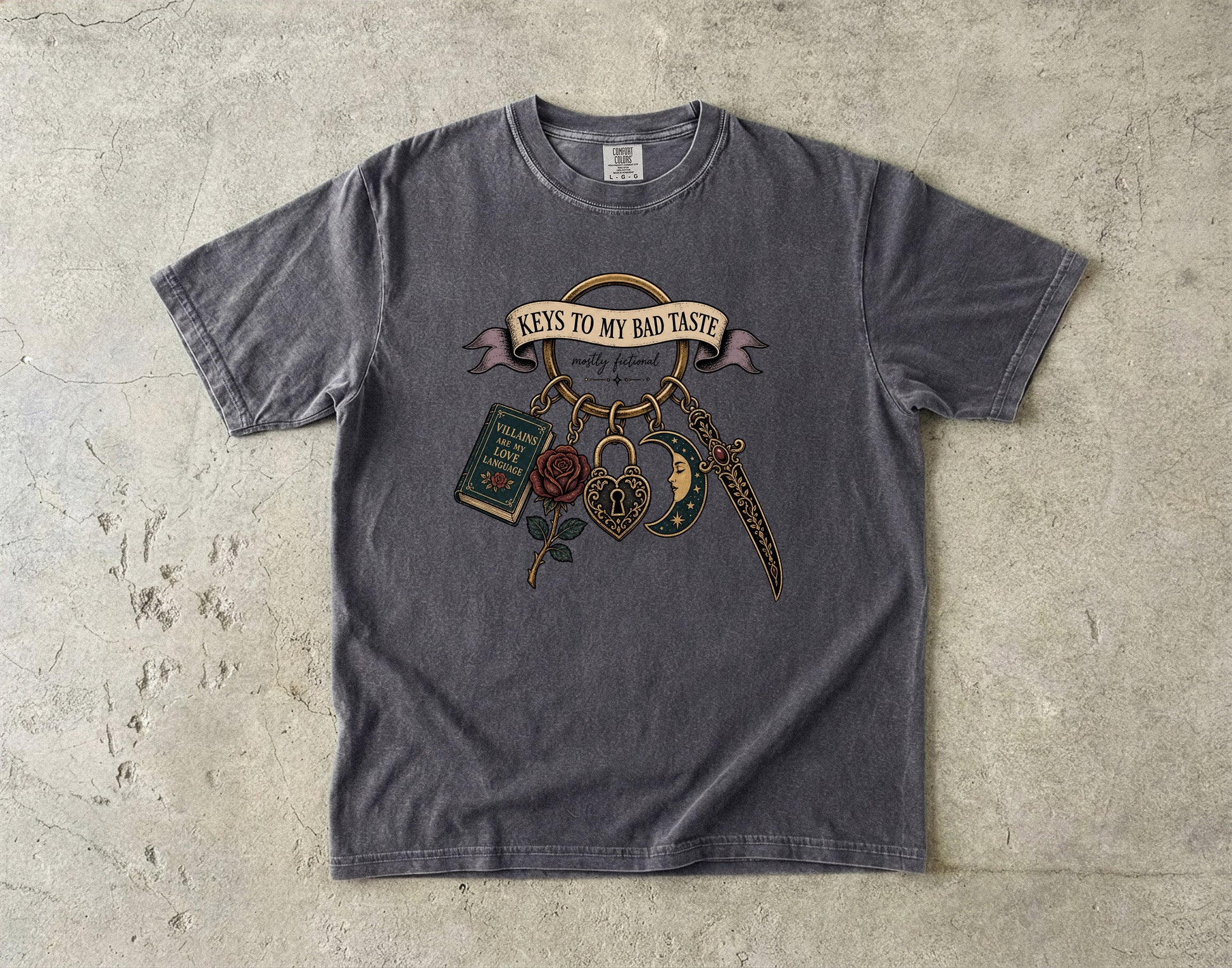A dark gray graphic t-shirt laid flat on a textured concrete surface. The shirt features a vintage-style tattoo illustration with the text 'KEYS TO MY BAD TASTE' on a banner, accompanied by a dagger, padlock, book, rose, and keychain.