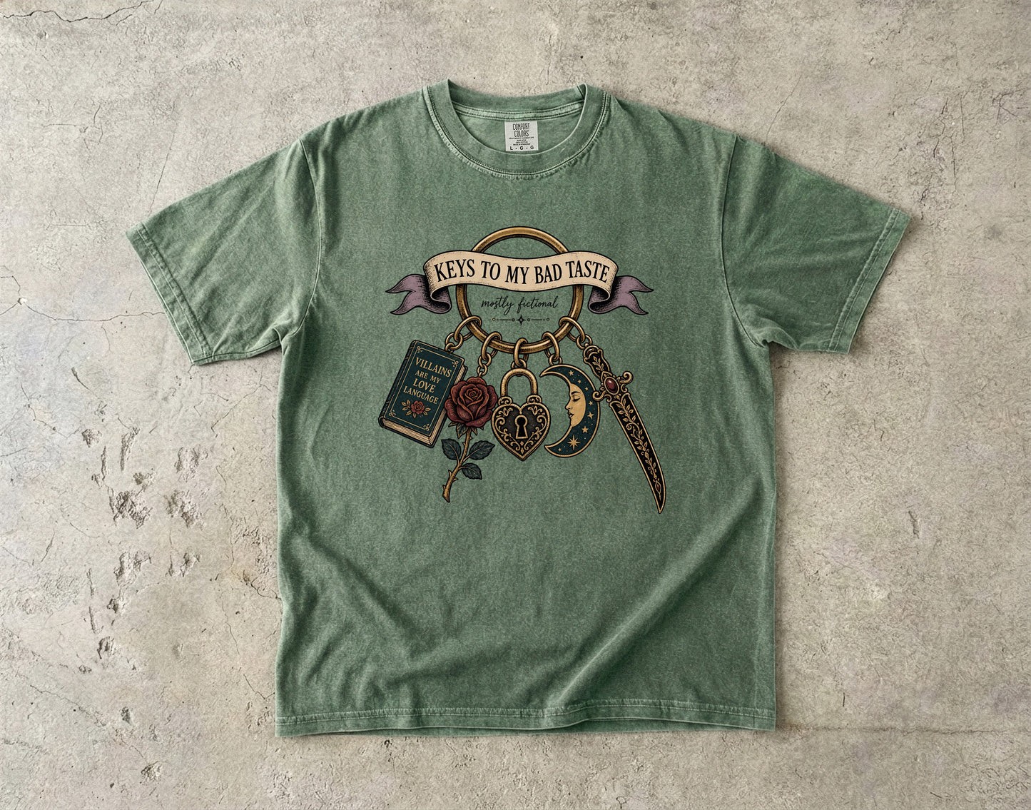 A sage green short-sleeve t-shirt with a vintage-style graphic on the front, laid flat on a textured concrete surface. The graphic features a banner reading 'KEYS TO MY BAD TASTE' with hanging illustrations of a book, rose, dagger, and padlock.