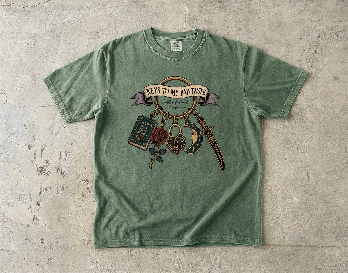 A sage green short-sleeve t-shirt with a vintage-style graphic on the front, laid flat on a textured concrete surface. The graphic features a banner reading 'KEYS TO MY BAD TASTE' with hanging illustrations of a book, rose, dagger, and padlock.