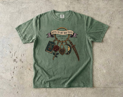 A sage green short-sleeve t-shirt with a vintage-style graphic on the front, laid flat on a textured concrete surface. The graphic features a banner reading 'KEYS TO MY BAD TASTE' with hanging illustrations of a book, rose, dagger, and padlock.