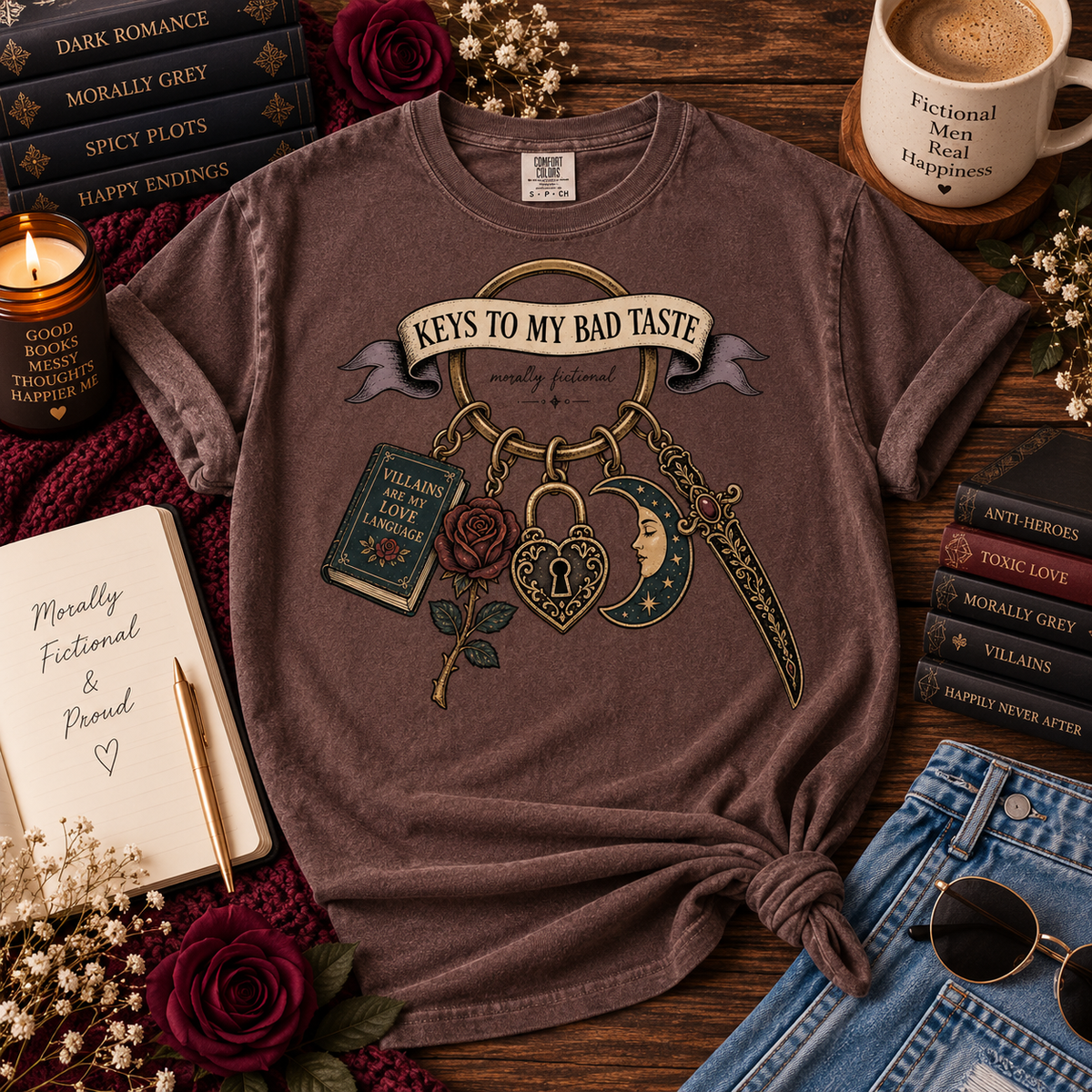 A graphic t-shirt in a dusty mauve-brown color, styled in a cozy flat-lay arrangement with books, a mug, sunglasses, and flowers on a wooden table. The shirt features a graphic of a keychain with a rose, moon, and book, and the text 'KEYS TO MY BAD TASTE'.