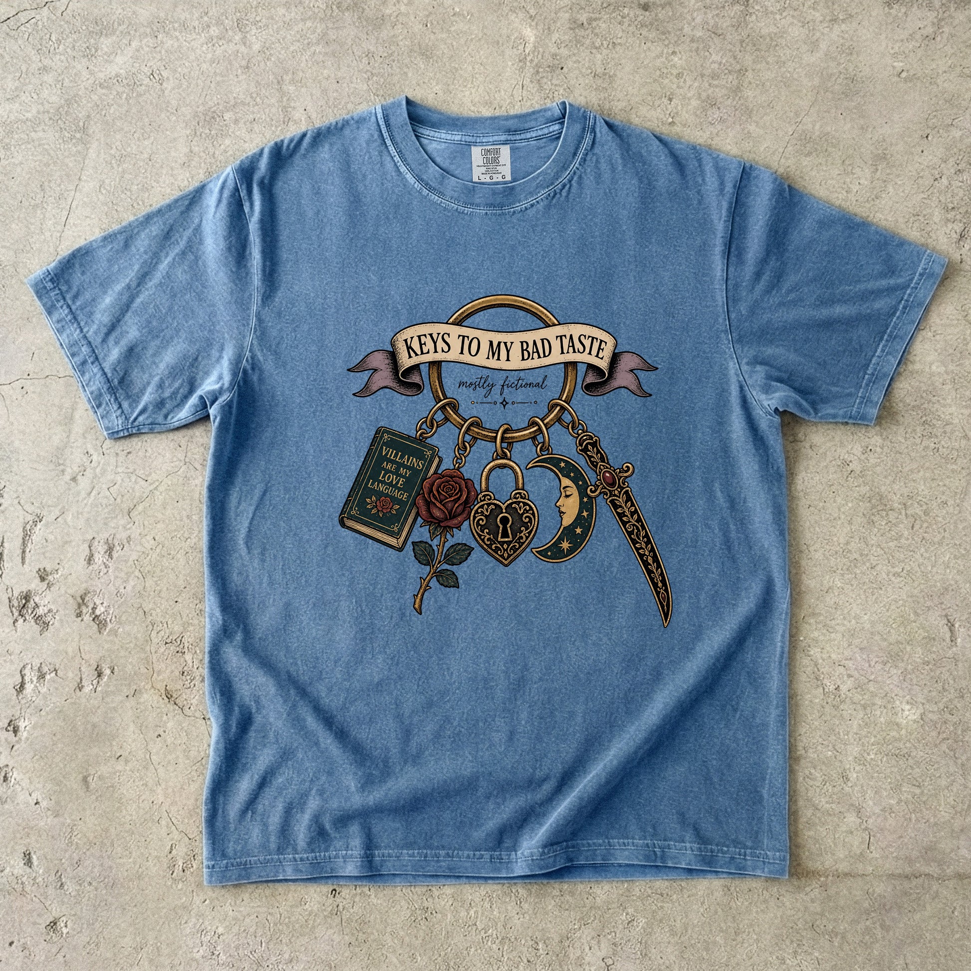 A light blue short-sleeve graphic t-shirt laid flat on a textured concrete surface. The graphic features a vintage tattoo-style design with the text 'KEYS TO MY BAD TASTE' on a banner, accompanied by a rose, padlock, dagger, and book charm.