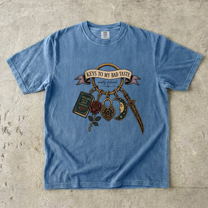 A light blue short-sleeve graphic t-shirt laid flat on a textured concrete surface. The graphic features a vintage tattoo-style design with the text 'KEYS TO MY BAD TASTE' on a banner, accompanied by a rose, padlock, dagger, and book charm.