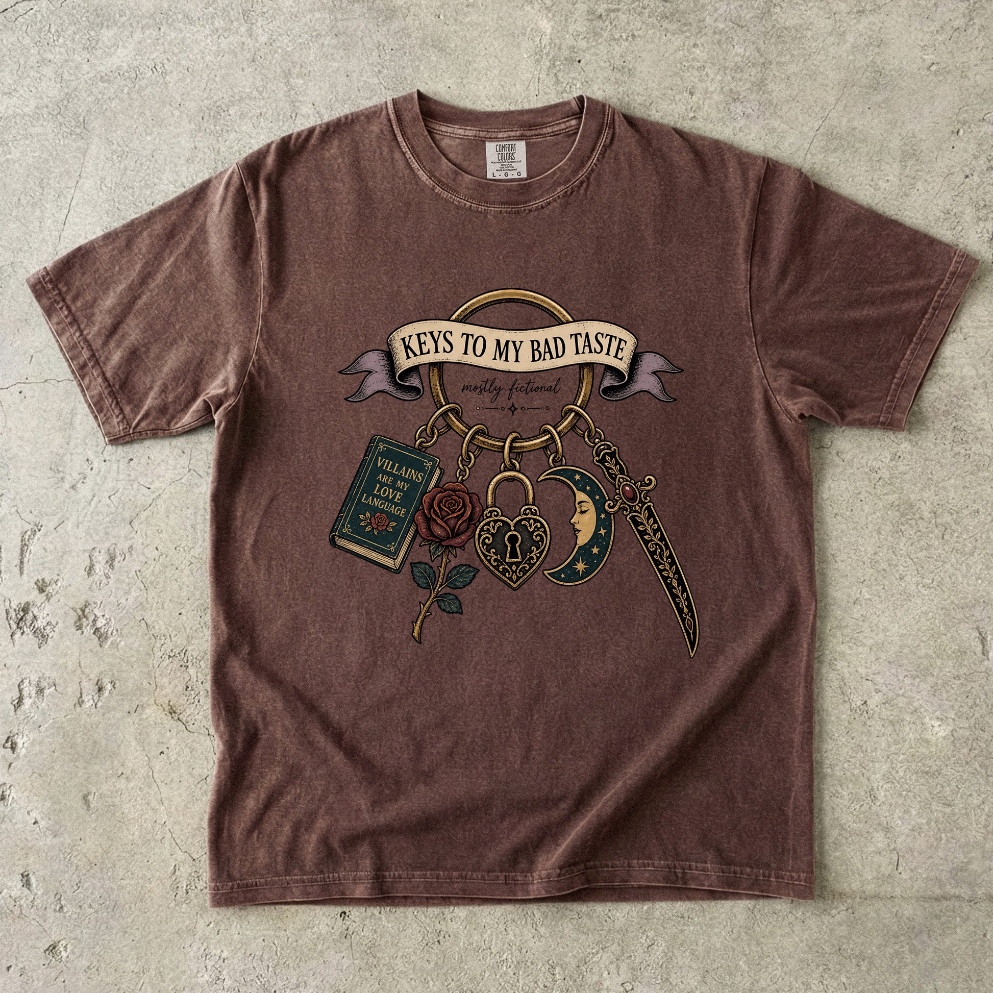 A brown short-sleeve t-shirt with a vintage-style graphic print featuring the text 'KEYS TO MY BAD TASTE' and illustrations of a book, rose, padlock, dagger, and crescent moon, laid flat on a textured concrete surface.