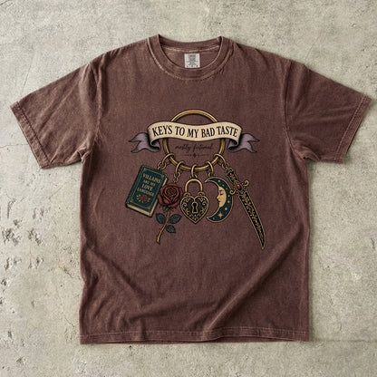 A brown short-sleeve t-shirt with a vintage-style graphic print featuring the text 'KEYS TO MY BAD TASTE' and illustrations of a book, rose, padlock, dagger, and crescent moon, laid flat on a textured concrete surface.