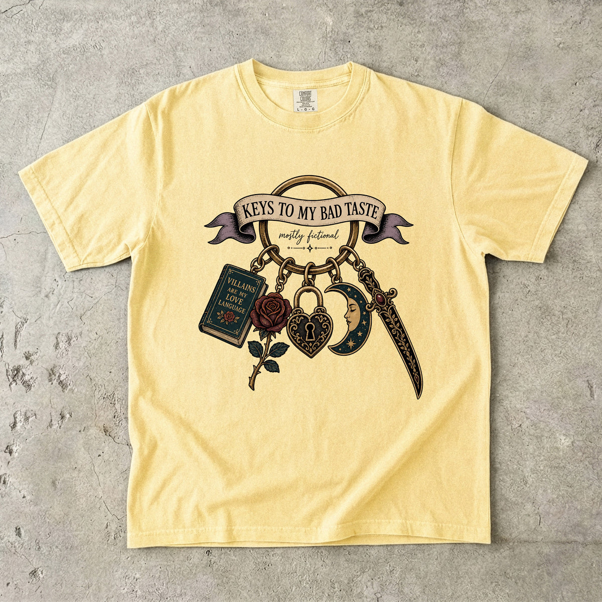 A pale yellow graphic t-shirt laid flat on a textured gray concrete surface. The shirt features a detailed graphic print of a keychain with a dagger, a rose, a book, and a heart-shaped padlock under a banner that reads 'KEYS TO MY BAD TASTE'.