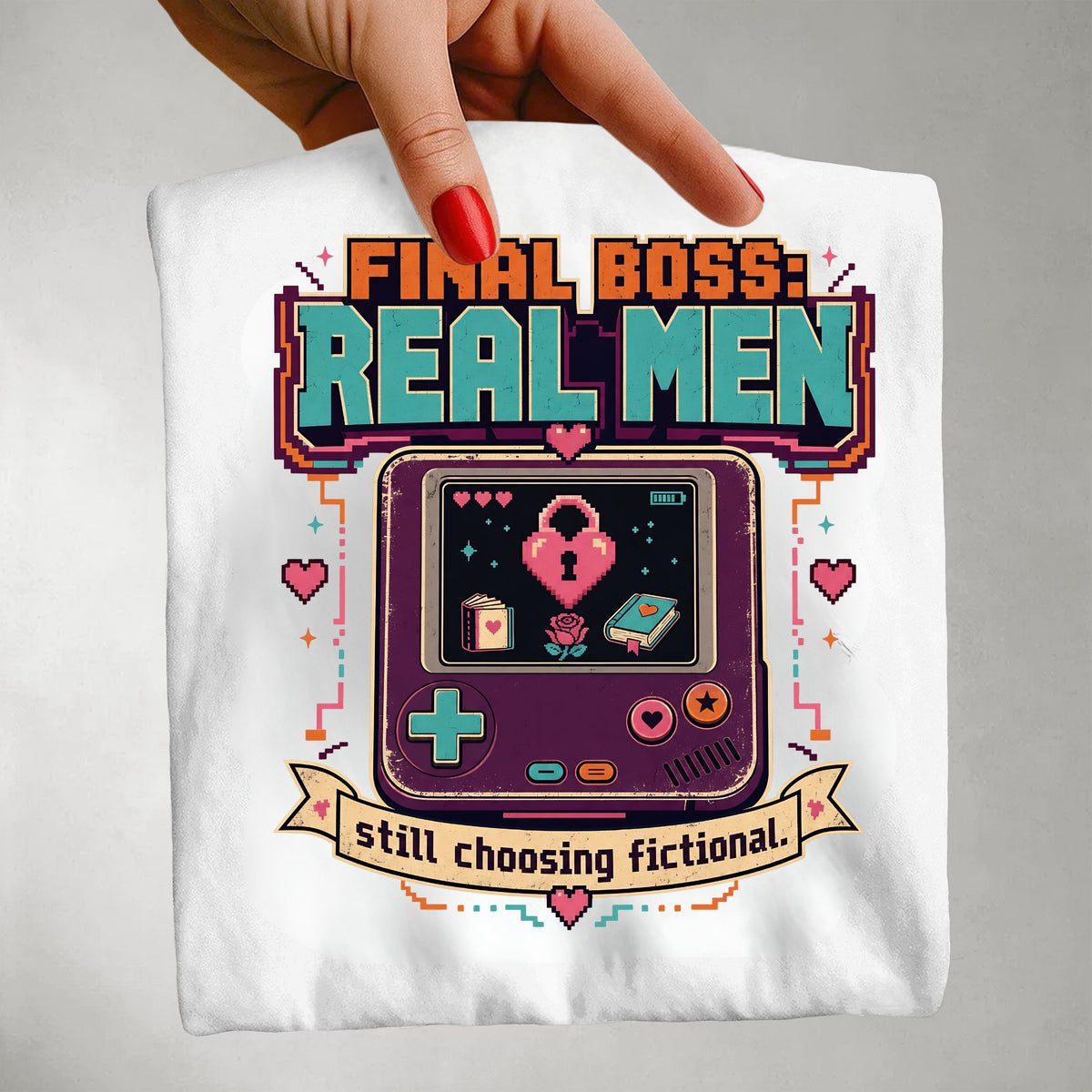 A hand with red nail polish holds up a white t-shirt featuring a colorful retro video game console graphic and the text 'FINAL BOSS: REAL MEN still choosing fictional.' against a neutral gray background.