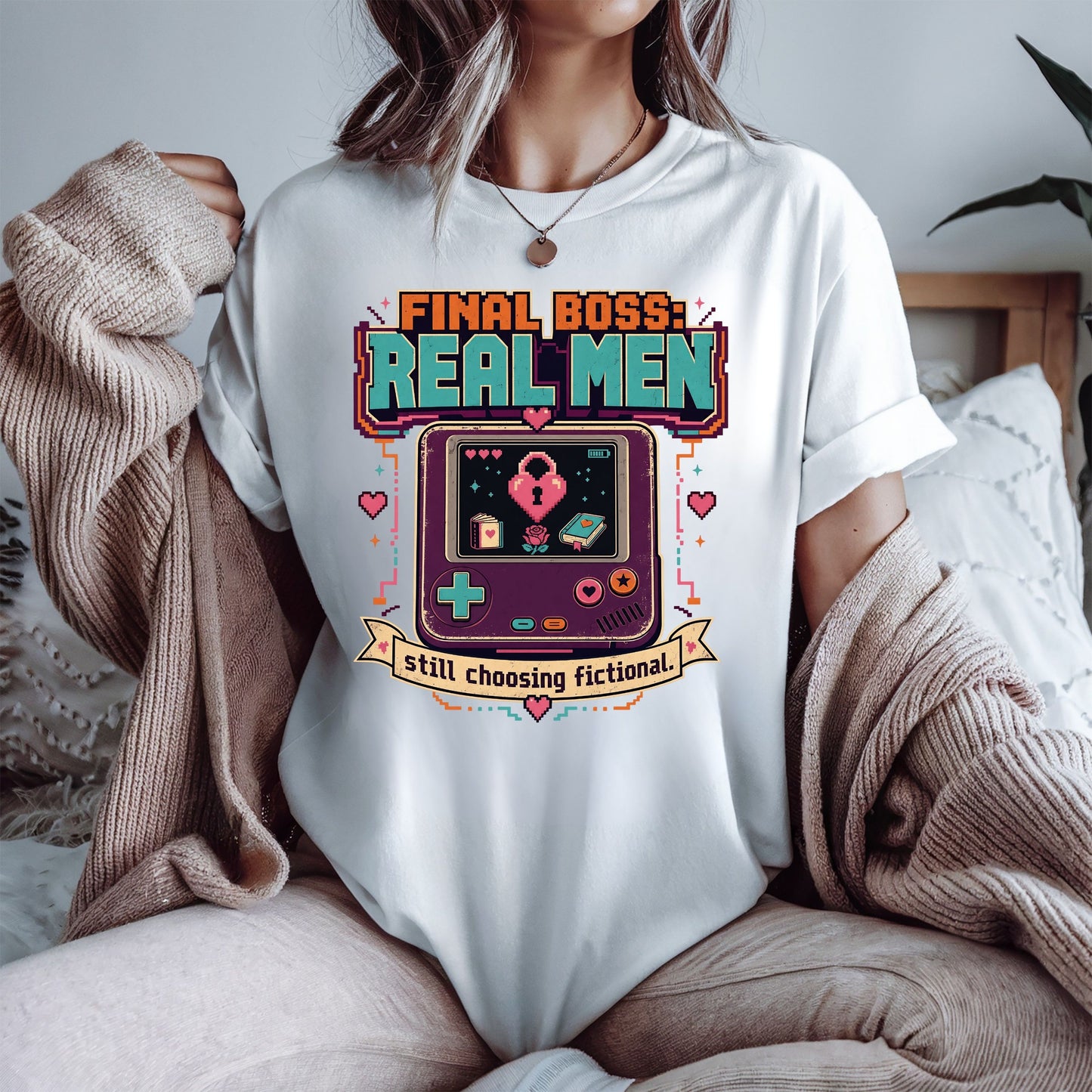 A woman wearing a white graphic t-shirt with a colorful retro video game console design and the text 'FINAL BOSS: REAL MEN' and 'still choosing fictional'. She is wearing a beige knit cardigan draped off her shoulders in a cozy indoor setting.